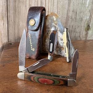 May include: A collection of pocket knives and a leather sheath. The knives have wooden and metal handles, with blades in various positions. The sheath is brown leather with the word "LEATHERMAN" printed on it.