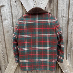 May include: A vintage plaid jacket in shades of green, red, and brown, with a brown faux fur collar. The jacket is hanging on a wooden background, showcasing its classic design and warm color palette. The plaid pattern is a traditional style.