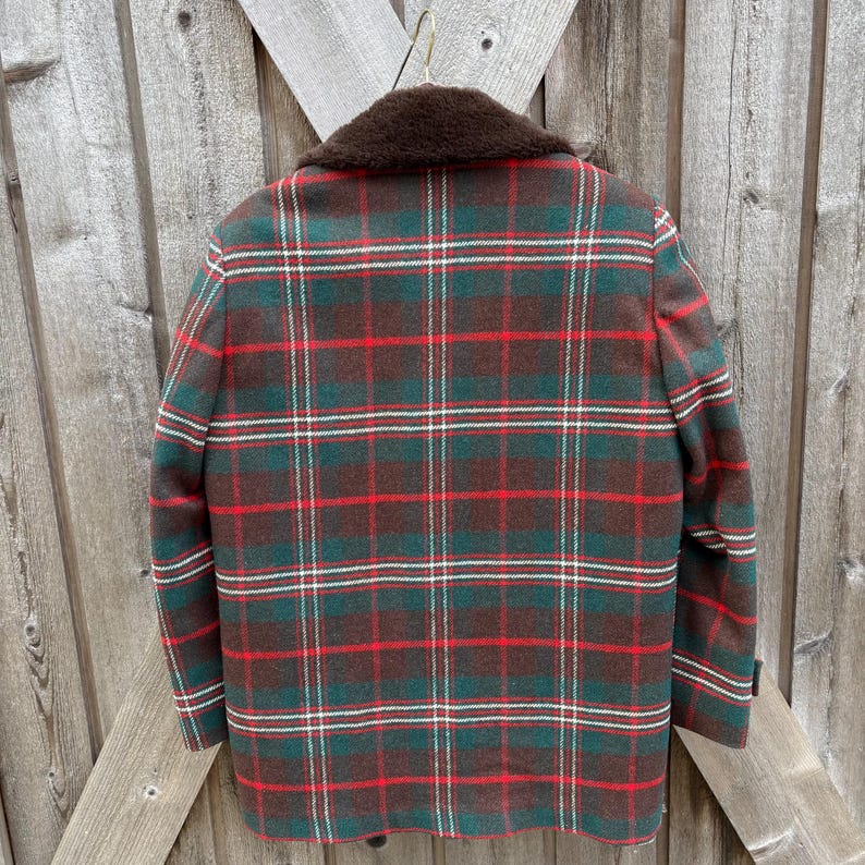 May include: A vintage plaid jacket in shades of green, red, and brown, with a brown faux fur collar. The jacket is hanging on a wooden background, showcasing its classic design and warm color palette. The plaid pattern is a traditional style.