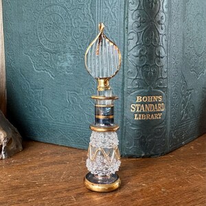May include: A decorative glass perfume bottle with a gold-colored base and accents. The bottle has a clear, textured body and a gold-trimmed, teardrop-shaped stopper. The bottle is approximately 5 inches tall.