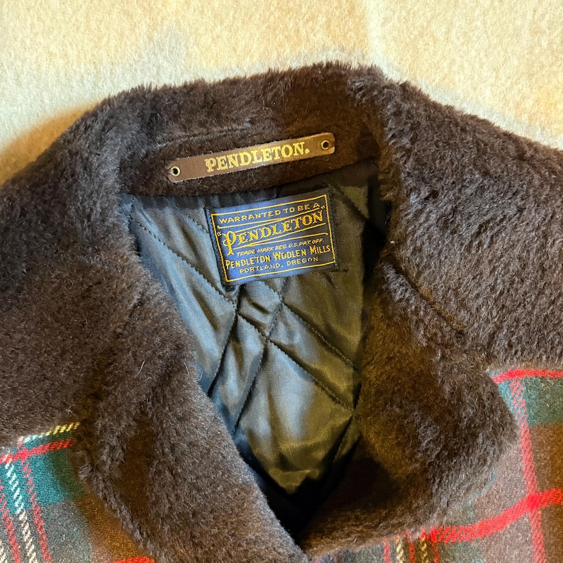 May include: Close-up of a Pendleton jacket collar with a brown fur-like trim. The jacket features a plaid pattern in red, green, and brown. A label reads "PENDLETON" and another label reads "WARRANTED TO BE A PENDLETON".
