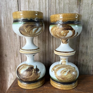 May include: Two ceramic goblets with a unique design. The goblets have a white base with brown swirl patterns and a brown rim. The top rim is a darker brown with a black band. The goblets are approximately 8 inches tall.