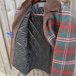 May include: A vintage plaid jacket with a brown faux fur collar and a black quilted lining. The jacket features a green, red, and white plaid pattern. The jacket is hanging on a wooden hanger.