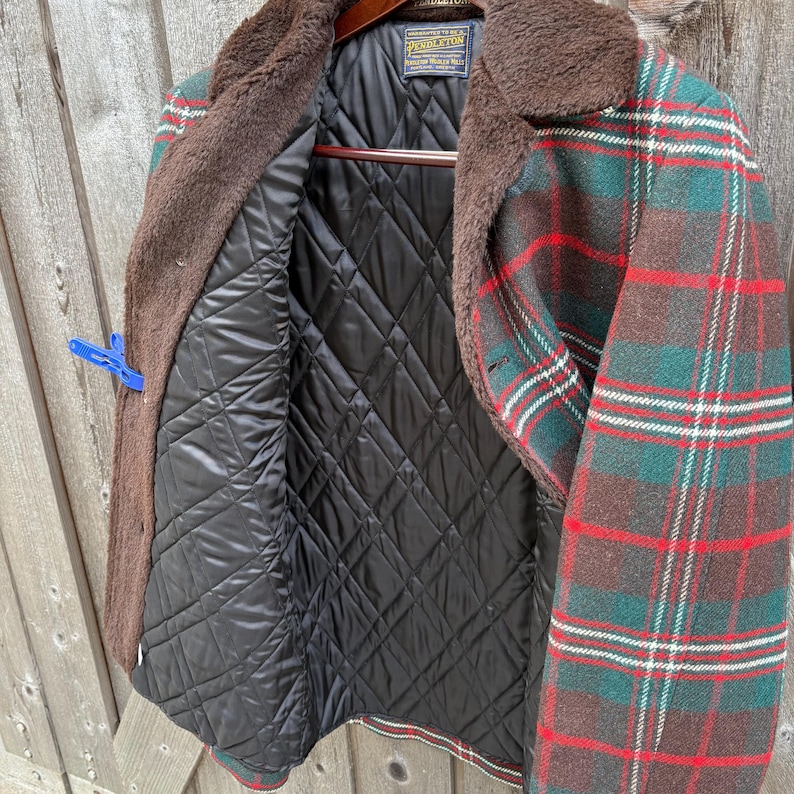 May include: A vintage plaid jacket with a brown faux fur collar and a black quilted lining. The jacket features a green, red, and white plaid pattern. The jacket is hanging on a wooden hanger.
