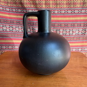 May include: A matte black ceramic pitcher with a rounded body and a handle. The pitcher sits on a wooden surface, with a patterned textile in the background. The pitcher is a decorative item.