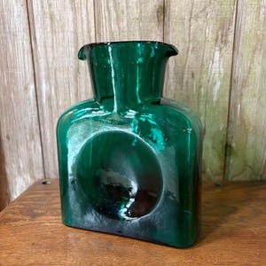 May include: A dark green glass vase with a unique, rounded design. The vase has a square base and a circular indentation on the front. The top has a flared, irregular shape. The glass has a glossy finish.