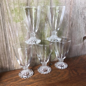May include: A set of six clear glass goblets. Each goblet has a flared bowl with vertical ribbing and sits atop a decorative base with a beaded design. The glasses are arranged on a wooden surface against a weathered wood background.