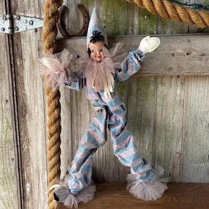 May include: A vintage clown doll with a blue and pink striped outfit, a ruffled collar, and a pointed hat. The doll has white gloves and tulle accents. The doll is posed with arms outstretched and legs apart.