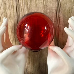 May include: A large, round, red glass ball. The ball is held by two white hands. The glass is reflective, with highlights. The background is a wooden surface.