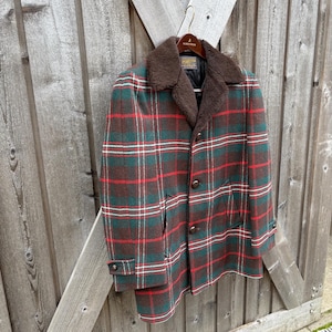 May include: A vintage plaid coat in shades of green, red, and brown, with a brown faux fur collar. The coat has a button-up front and is hanging on a wooden hanger against a wooden fence.