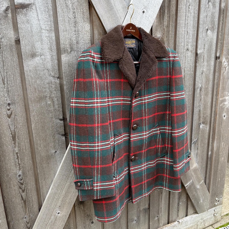 May include: A vintage plaid coat in shades of green, red, and brown, with a brown faux fur collar. The coat has a button-up front and is hanging on a wooden hanger against a wooden fence.