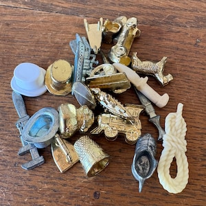 May include: A collection of assorted metal and plastic game tokens including a gold-tone top hat, a silver-tone ship, a gold-tone train, a thimble, a silver-tone shoe, a white plastic top hat, a white plastic rope, and a white plastic sword.