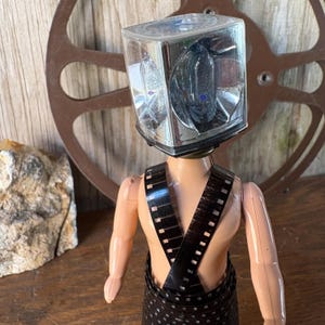 May include: A doll with a clear, square light fixture for a head and film strip wrapped around its torso. The doll is tan with a brown film reel in the background. The light fixture has a silver interior.