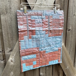 May include: A handmade quilt with a patchwork design. The quilt features a combination of red and blue fabric pieces, stitched together with dark thread. The quilt is hanging on a clear plastic hanger against a wooden background.