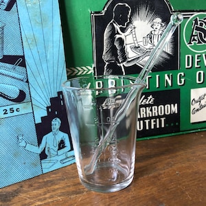 May include: A clear glass measuring cup with a pouring spout and measurements in ounces. A clear glass stirring rod rests inside the cup. A vintage blue and green box is in the background.