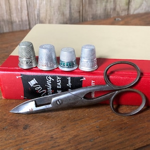 May include: Four metal thimbles with textured surfaces and a pair of vintage metal sewing scissors with the text J.Y. MOORE engraved on the handle. The scissors feature a pointed blade and two circular finger loops, resting on a red and beige book.