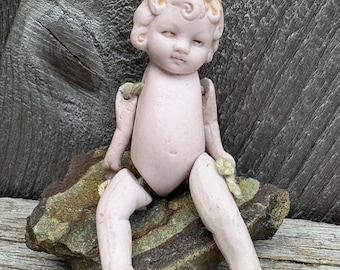 Könnte beinhalten: Eine Vintage-Puppenfigur aus Biskuitporzellan mit matter, blasser Oberfläche und modelliertem, hellorangem Haar. Die Puppe hat bewegliche Gliedmaßen, die mit sichtbaren beigen Schnüren verbunden sind, einen neutralen Gesichtsausdruck und sitzt.