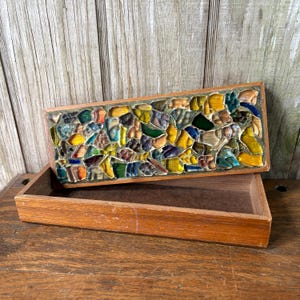 May include: A rectangular wooden box with a mosaic lid. The mosaic is composed of irregularly shaped pieces in shades of yellow, green, blue, and brown. The box is open, revealing a plain wooden interior. The background is a weathered wooden surface.