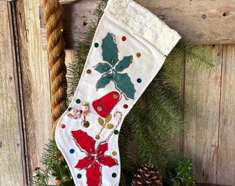 May include: A white fabric Christmas stocking featuring a plush cuff, decorated with green and red felt poinsettia appliqués, a red bell, small snowman figures, and scattered colorful sequins. The stocking is accented with gold stitching.