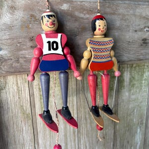 May include: Two vintage wooden marionette dolls. One doll has a red and white top with the number 10, blue shorts, and gray legs. The other doll has a red and blue top with a patterned design and red legs. Both dolls have skis and are hanging.