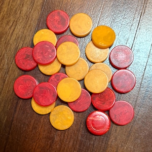 May include: A collection of red and yellow game pieces, likely for a board game. The circular pieces are scattered on a wooden surface. The red pieces have a marbled appearance, while the yellow pieces are solid in color.