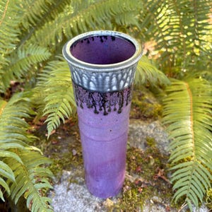 May include: A tall cylindrical ceramic vase featuring a vibrant purple matte finish on the lower body. The upper rim is glazed in a contrasting metallic silver with black drip patterns cascading down toward the purple section of the vessel.