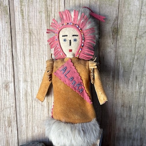 May include: A handmade doll featuring a tan suede-like body, adorned with a pink headdress and a sash that reads "ALASKA." The doll has a white face with blue eyes and a red mouth. The bottom of the doll is trimmed with white fur.