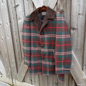 May include: A vintage plaid coat in shades of green, red, and brown. The coat features a brown faux fur collar, button closures, and pockets. The coat is hanging on a wooden hanger against a wooden fence.