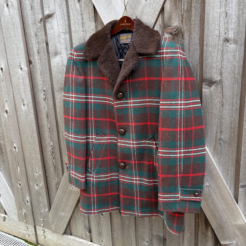 May include: A vintage plaid coat in shades of green, red, and brown. The coat features a brown faux fur collar, button closures, and pockets. The coat is hanging on a wooden hanger against a wooden fence.