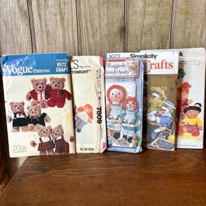 May include: A collection of vintage craft pattern books. The patterns include teddy bears, Raggedy Ann & Andy dolls, and other soft toys. The books are in various sizes and colors, with text and illustrations on the covers. The Vogue Patterns and Simplicity brands are visible.