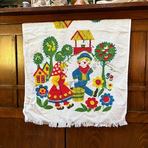 May include: White kitchen towel with a colorful, vintage design. The towel features a whimsical scene with cartoon figures, houses, trees, and flowers in red, yellow, blue, and green. The bottom edge has a fringe.