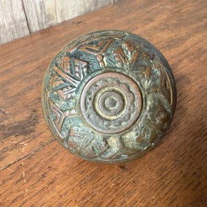May include: Antique bronze door knob with an intricate, raised design. The knob has a circular shape with a central, concentric circle pattern. The bronze has a weathered, aged appearance with hints of green and brown.