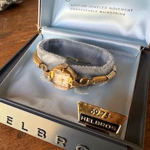 May include: A vintage gold-toned wristwatch with a rectangular face, resting in a blue velvet-lined box. The watch has a delicate, linked band. The box also contains a gold-colored pin with the text "69.75 HELBROS". The box reads "ELBRO".
