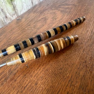 May include: Two vintage serving utensils with handles featuring alternating bands of black, tan, and cream-colored materials. The utensils are set against a wooden surface, showcasing their decorative design and craftsmanship.