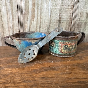 May include: An antique metal slotted spoon rests between two vintage tin cups. One cup is blue, the other is green with a label. The items show signs of age and wear, with rust and faded paint.
