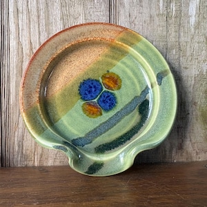 May include: A handmade ceramic spoon rest with a unique design. The spoon rest features a blend of colors, including green, orange, and blue. The center has a cluster of blue and orange floral accents. The spoon rest has a curved shape with a built-in rest.