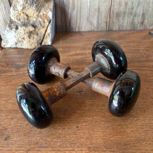 May include: Two vintage door knobs featuring smooth, glossy black spherical handles attached to weathered, rusted metal shanks and square spindles. The two units are crossed over each other, showcasing the contrast between the dark, reflective surfaces and the oxidized, textured metal components of the hardware.