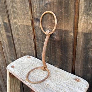 May include: A rusted metal horse bit featuring a curved central bar connected to two circular rings at each end. The iron surface displays a weathered, oxidized brown texture throughout the entire structure, showing signs of age and consistent wear.