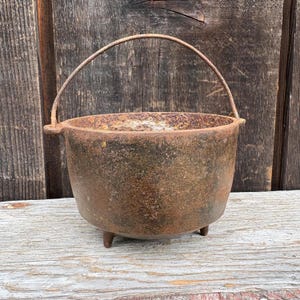 May include: A round cast iron cauldron with a heavy, weathered, rusted brown patina. The pot features three short, sturdy legs at the base and a curved, pivoting metal handle attached to two side lugs. The interior shows signs of oxidation.