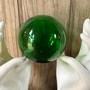 May include: A large, round, emerald green glass ball. The sphere reflects light, creating bright highlights. The ball is held by two white, sculpted hands against a wooden background. The overall theme is decorative.