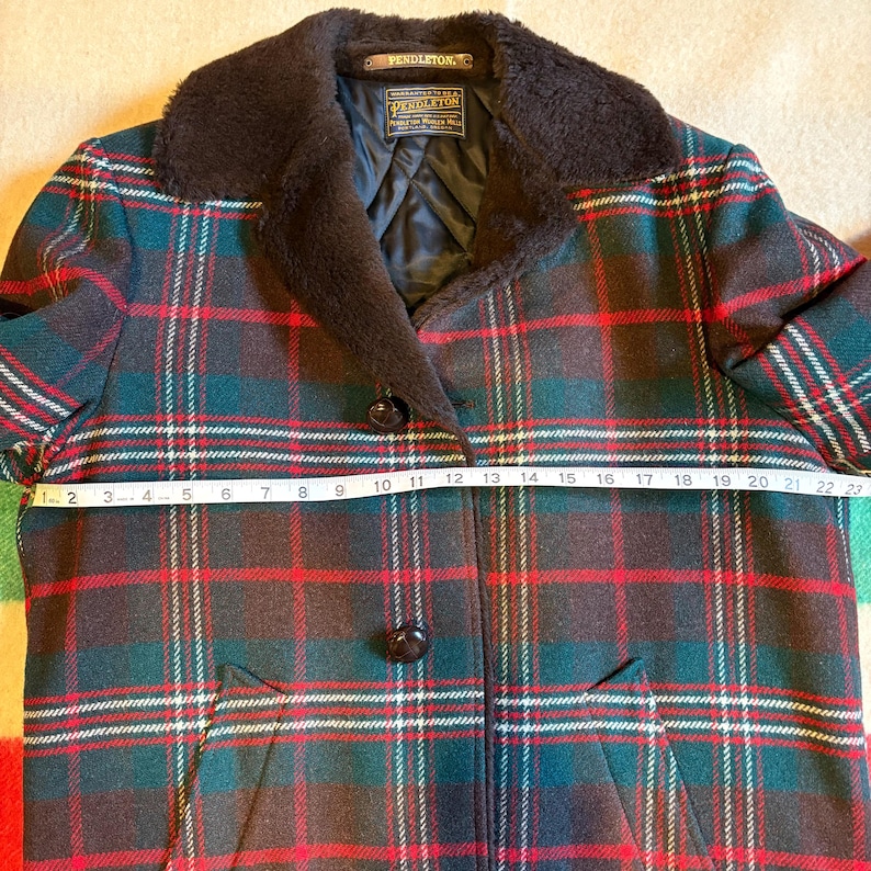 May include: A vintage Pendleton plaid coat with a dark brown faux fur collar. The coat features a classic red, green, and white plaid pattern. The coat measures approximately 23 inches across the chest.