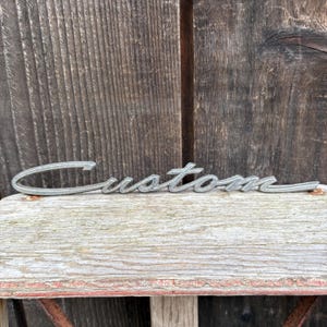 May include: A silver-toned metal emblem featuring the word Custom in a cursive, flowing script font. The lettering has a metallic finish with a slightly raised, three-dimensional appearance, designed for attachment to a vehicle or surface.