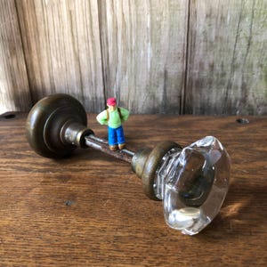 May include: Antique brass doorknob with a clear, faceted glass handle. The doorknob is set against a rustic wooden background. A miniature figure stands on the doorknob's rod, adding a whimsical touch. The doorknob is a vintage item.