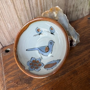May include: A small, round ceramic bowl with a light blue interior and a brown rim. The bowl is decorated with a blue bird, flowers, and butterflies. The bowl is likely handmade and has a rustic, folk art style.
