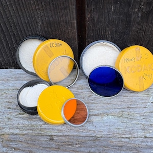 May include: A collection of vintage Kodak camera filters and accessories. The filters are in yellow and black cases, with clear, blue, and orange filter lenses. Some cases have text like "CC30M" and "KODAK C5".