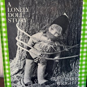 May include: A black and white photograph of a doll tied to a tree. The doll is wearing a black hat, a bandana, and a fringed jacket. The book cover reads "A Lonely Doll Story" by Dare Wright.