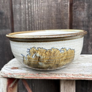 May include: A handmade ceramic bowl with a wide, shallow design. The bowl features a light beige base with a brown rim and a large, abstract brown and gold design on the side. The bowl is sitting on a weathered wooden surface.