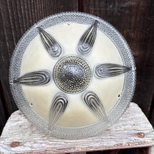 May include: A vintage, round ceiling light fixture with a cream-colored center and a silver-colored, beaded edge. The center features a textured pattern and a central, circular element. Six clear, teardrop-shaped elements radiate from the center.