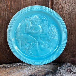May include: A round, light blue ceramic plate featuring a raised relief design of a person sitting at a pottery wheel, shaping a vessel. The scene includes a potted plant and architectural elements in the background, with a beaded border rim.