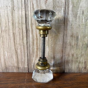 May include: Clear glass door knob with a ribbed top and a faceted bottom. The knob has brass-colored metal accents and a threaded rod. The background is a wooden surface.